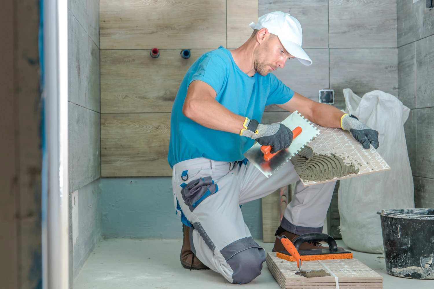 Hombre instalando azulejos en un baño, usando una llana para aplicar mortero en una pieza de cerámica. El fondo muestra paredes de azulejos de madera y herramientas de trabajo.
