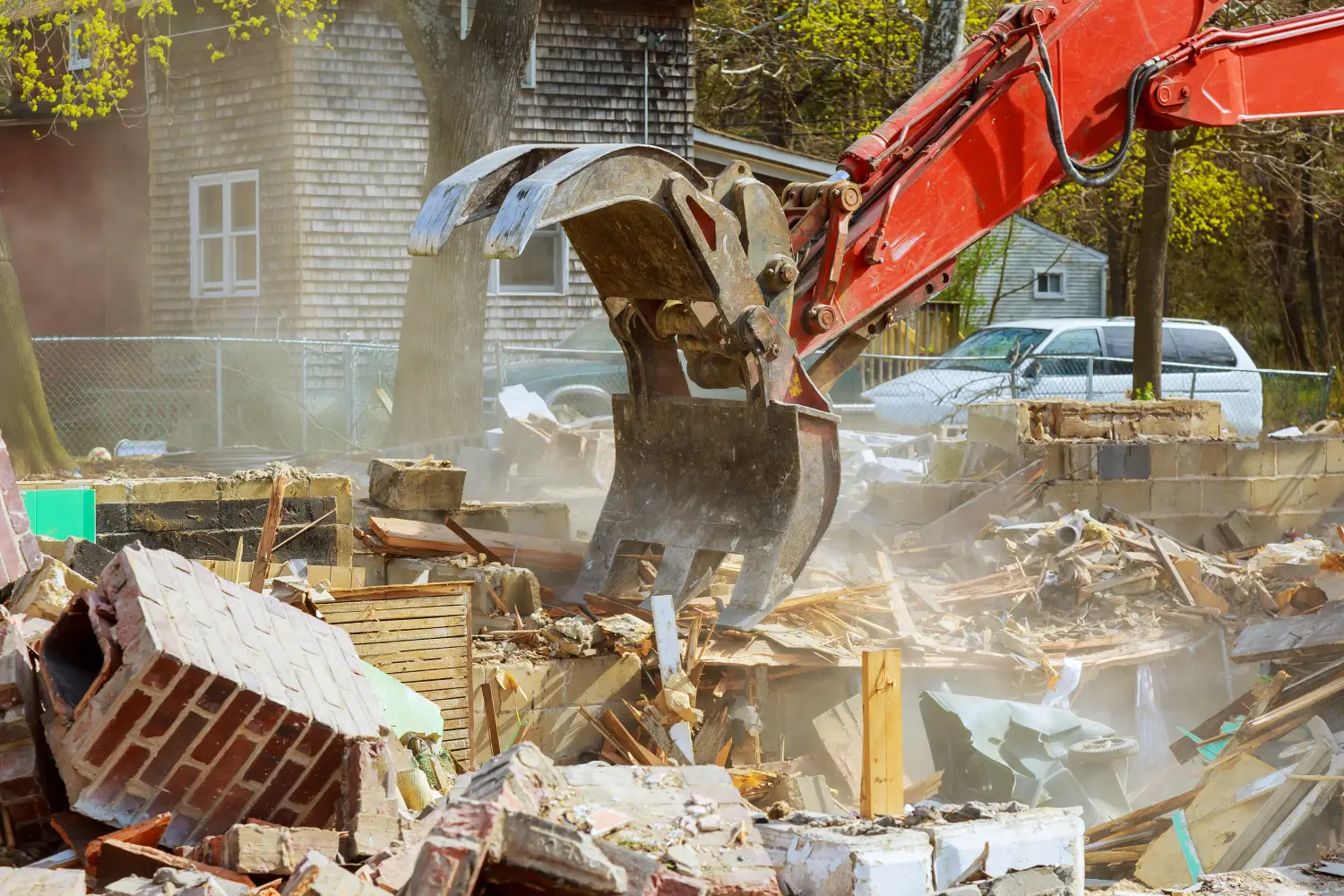 Excavadora roja levantando escombros en un sitio de demolición, con polvo en el aire y restos de materiales de construcción visibles.