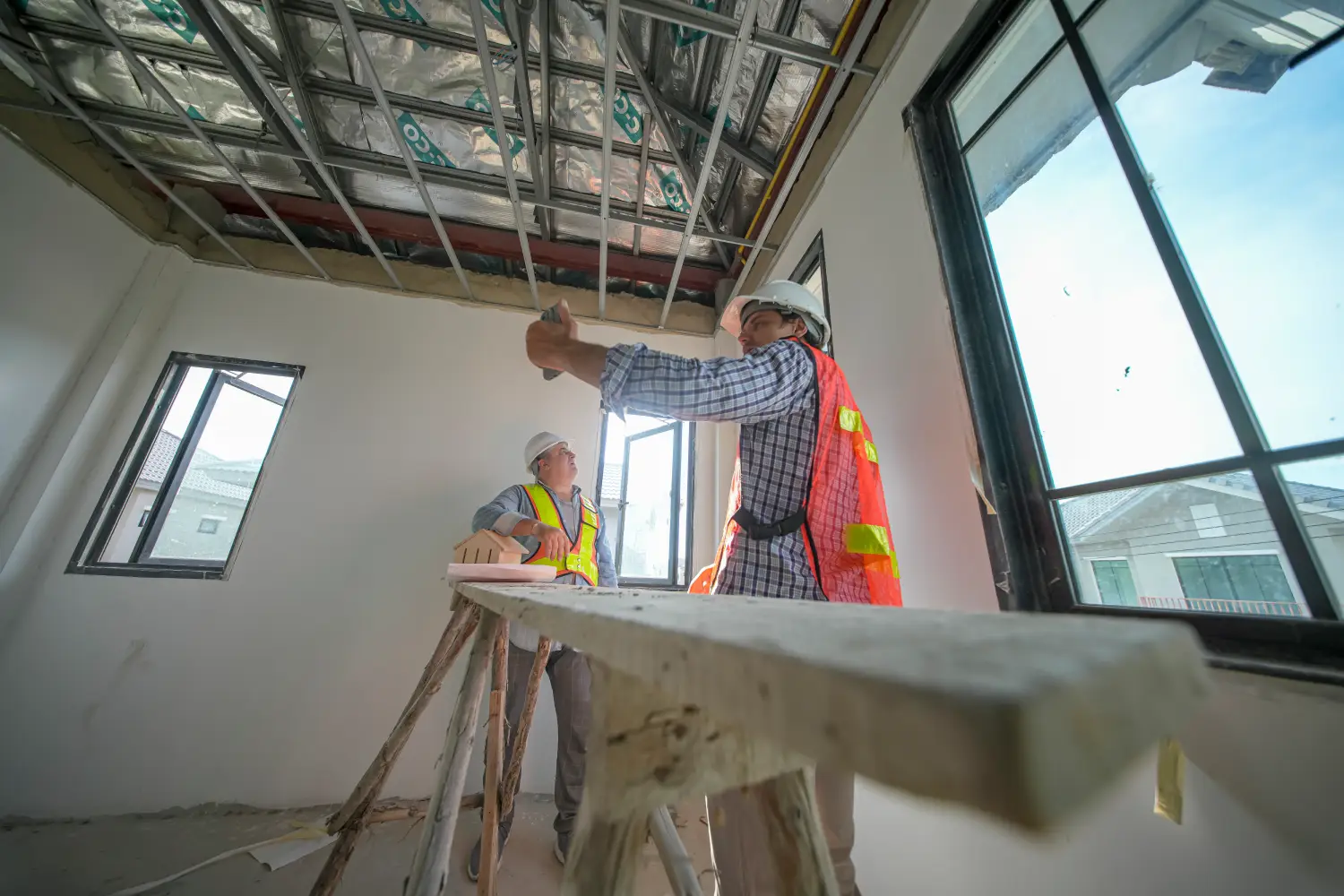 Dos trabajadores de la construcción en un interior en proceso de remodelación, usando cascos y chalecos de seguridad. Uno de ellos sostiene una herramienta mientras el otro observa. La imagen muestra vigas expuestas y una ventana con luz natural.