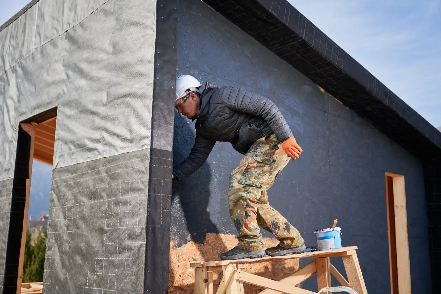 Trabajador de la construcción aplicando revestimiento en la pared de una casa en construcción, usando casco y guantes, mientras se encuentra sobre una plataforma de madera.